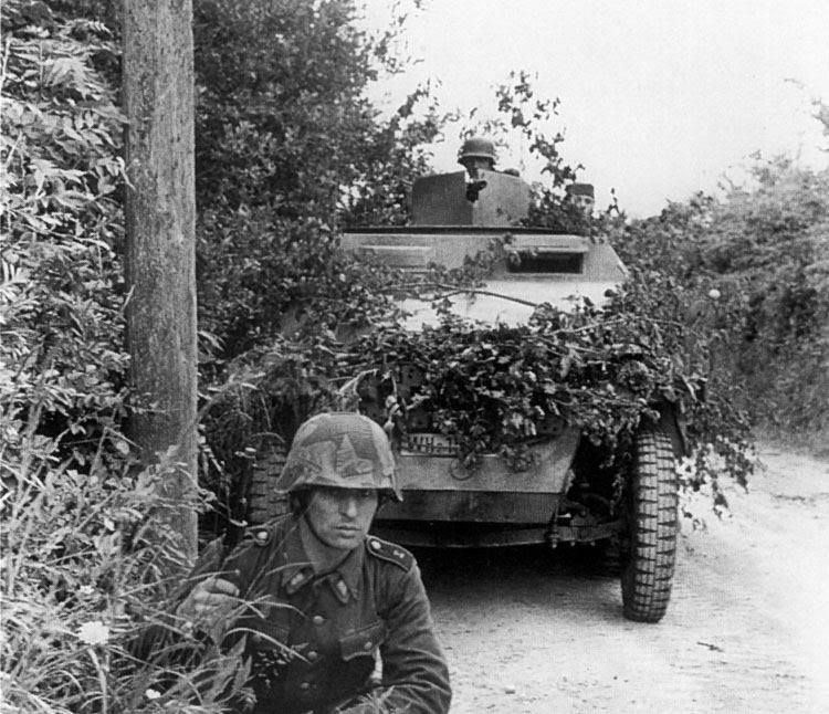 A grenadier crouching by a hedgerow with a camouflaged Panzer Lehr armoured vehicle behind him